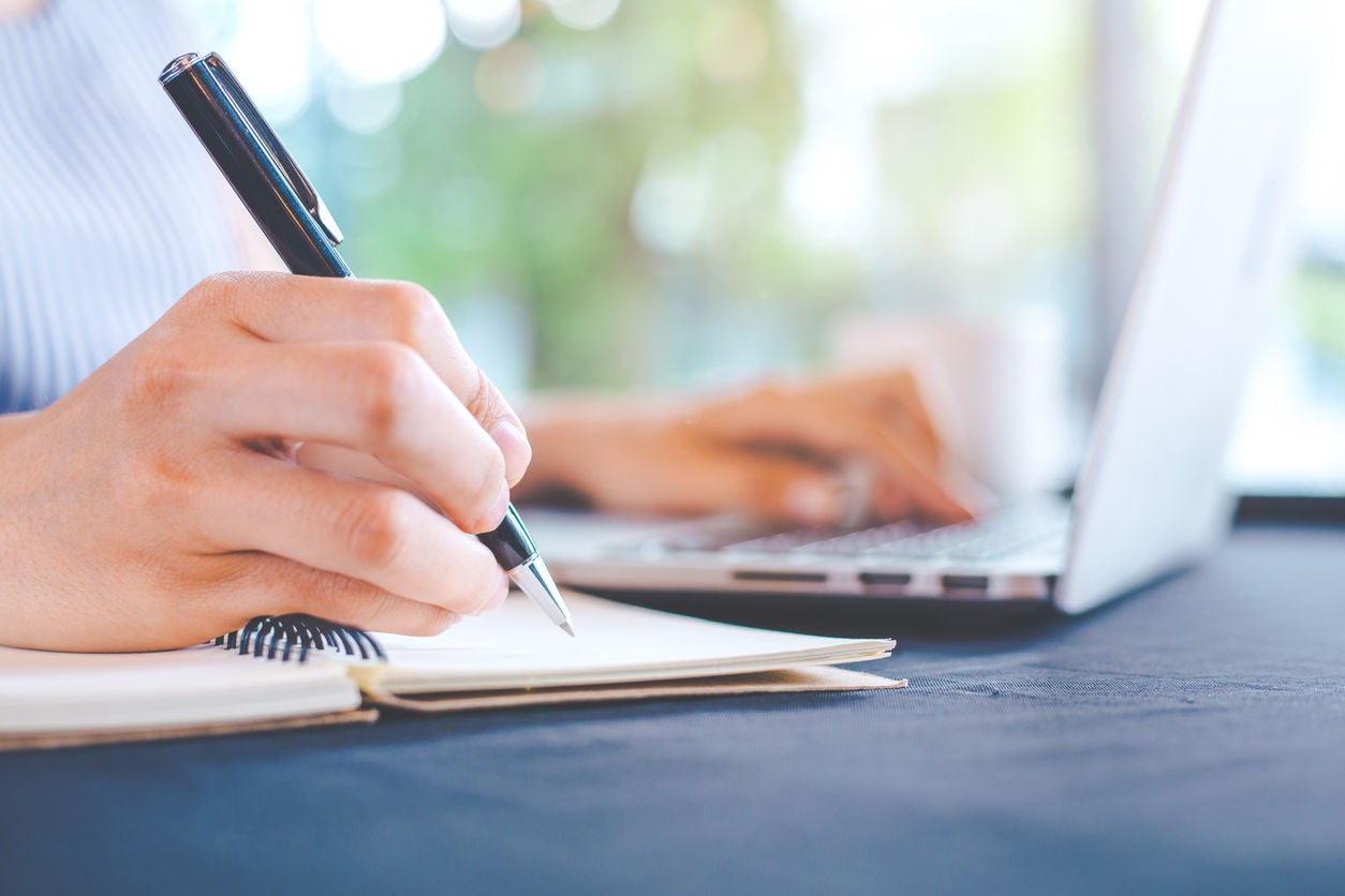 Woman hand writing on notepad with a pen and works in a laptop computer in the office.